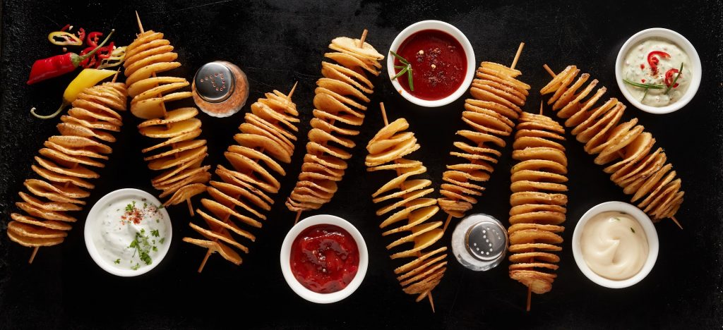 A top view of tasty fried tornado potatoes on sticks on a black table with various types of sauces